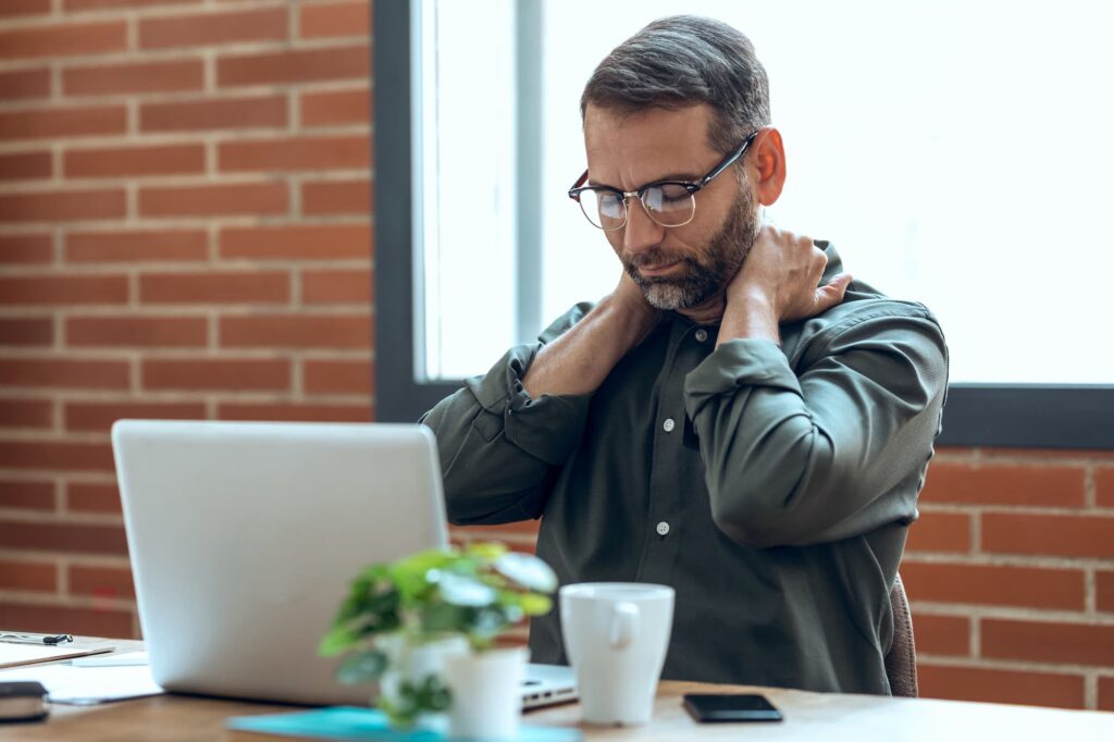 A middle-aged man sitting at a desk with a laptop, rubbing his neck and shoulders with a grimace on his face because he hasn't done neck exercises for pain near Bucktown.